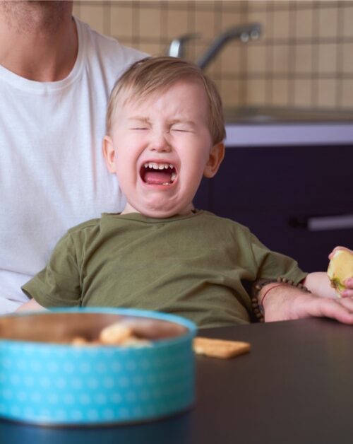 A small child is crying with a parent holding him in front of a blue bowl on a table